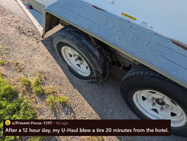 A completely shredded and blown-out tire on a white U-Haul trailer parked on a gravel shoulder.