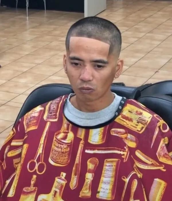 Man in a barber cape looking down with a sharp, exaggerated 90-degree rectangular hairline cut deep into his forehead, a painful haircut fail.