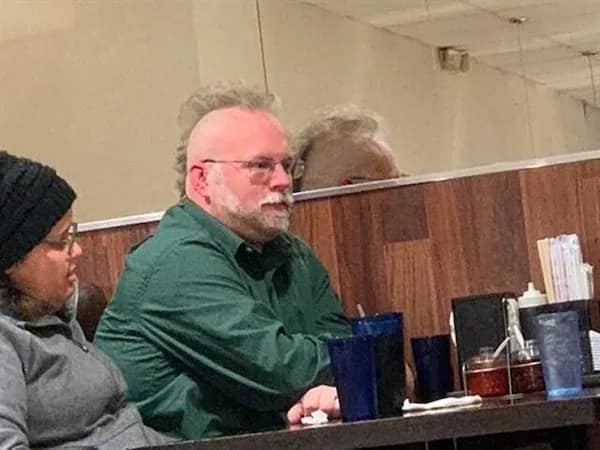 Older bearded man in a restaurant booth with shaved sides and a fluffy white mohawk strip, illustrating unique bad haircuts.
