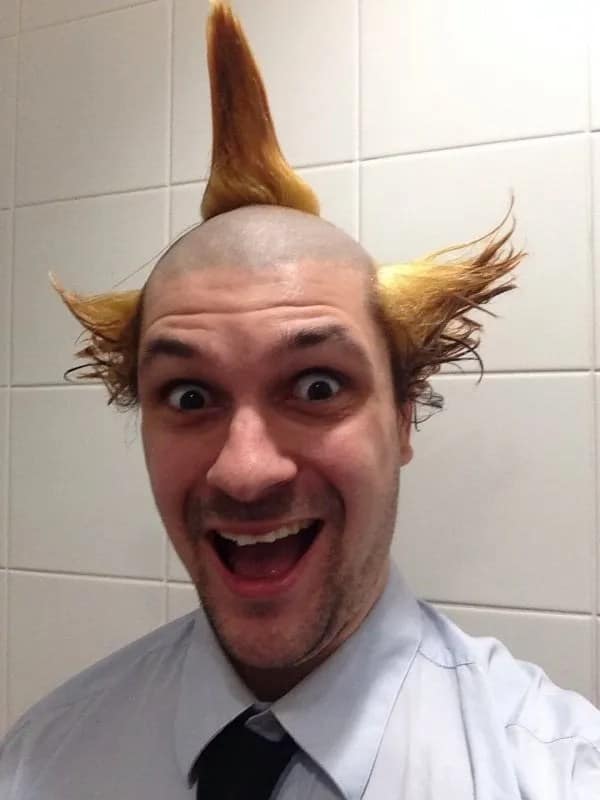 Close-up selfie of a smiling man with a shaved head styled into three pointed yellow hair spikes, a wild haircut fail.