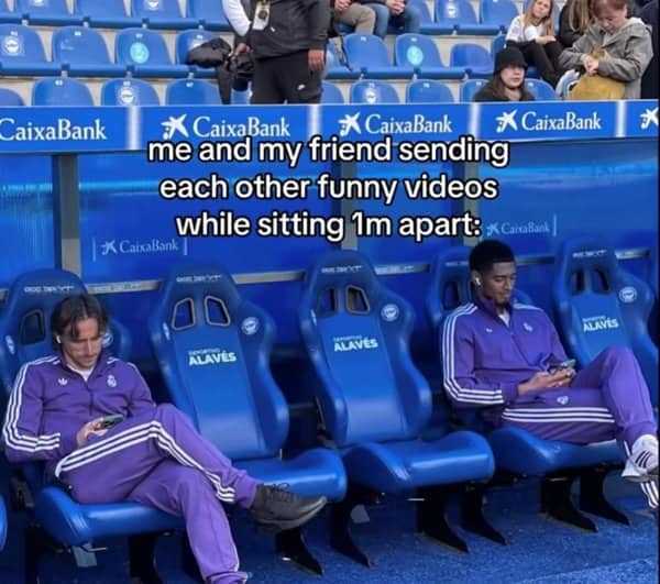 Two professional football players sitting far apart on stadium benches while looking at smartphones.