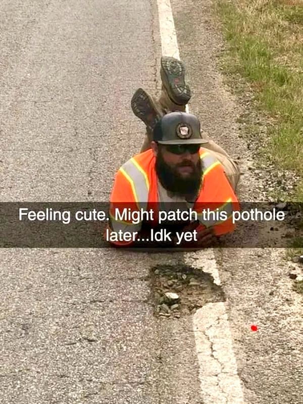 Construction worker in a high-visibility orange vest playfully posing on his stomach next to a large pothole.