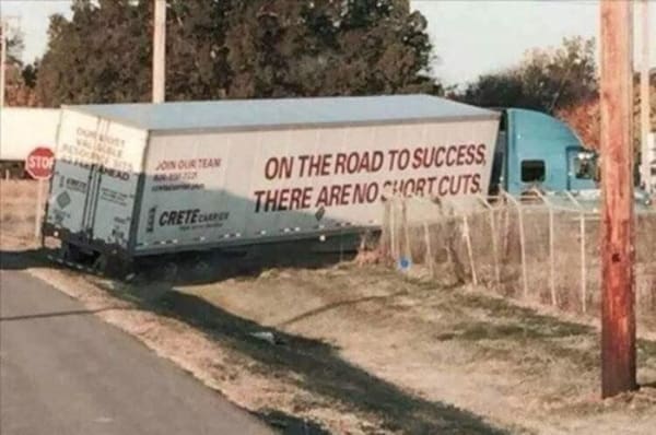 Large delivery semi-truck stuck in a roadside ditch directly underneath a printed slogan about taking no shortcuts.