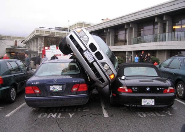A multi-vehicle parking lot collision serving as a spectacular car fail where a silver BMW sedan is wedged diagonally on its side across a blue Mercedes and a black BMW Z3 convertible in a space marked "Members Only".
