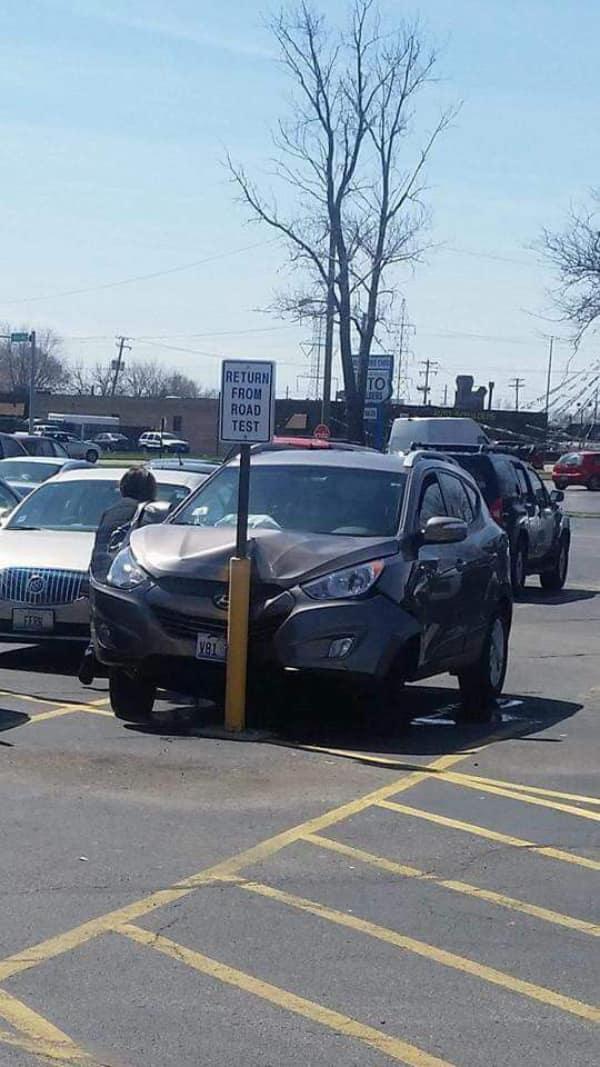 An ironic driver's evaluation car fail showing a silver SUV that has crashed directly center-front into a yellow parking lot barrier pole situated immediately below a "RETURN FROM ROAD TEST" sign.