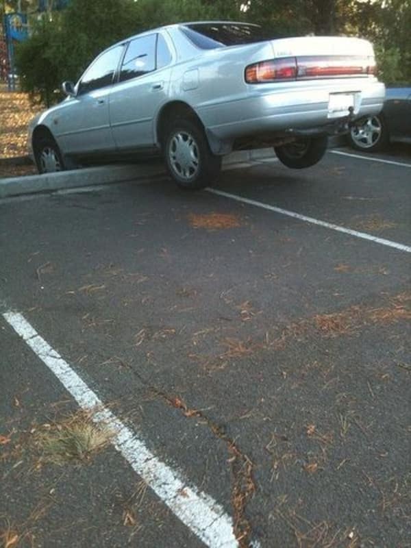 A high-centered parking lot car fail captures an older model silver sedan balanced precariously on top of a concrete curb block, leaving its rear wheels entirely suspended in mid-air over the empty space behind it.