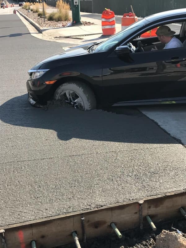 A standard road construction zone car fail captures a black sedan completely trapped and sunken front-wheel deep inside a freshly poured lane of wet, gray concrete.