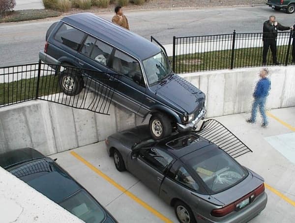 A chaotic multi-level parking structure car fail features an older dark blue SUV that has smashed through a black metal security fence, dangling off a high retaining wall to rest its front tire on top of a grey sports coupe below.