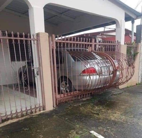 A bizarre property modification car fail showcases a silver sedan parked inside a closed carport, where the rear bumper protrudes onto the sidewalk through a wrought-iron gate custom-welded into a giant bubble cage to fit the vehicle.