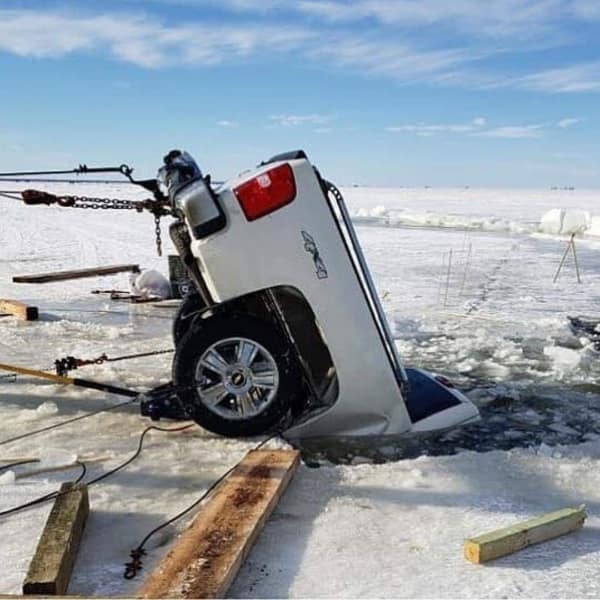 A winter recreation recovery car fail captures a white 4x4 pickup truck that has broken through melting lake ice rear-first, leaving its front nose pointed straight up at the sky while recovery winches try to salvage it.