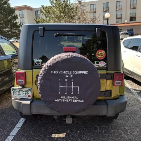 A close-up on the rear spare tire cover of a yellow Jeep Wrangler depicting a 6-speed manual stick shift pattern labeled as a "Millennial Anti-Theft Device" in a funny car fail trope.