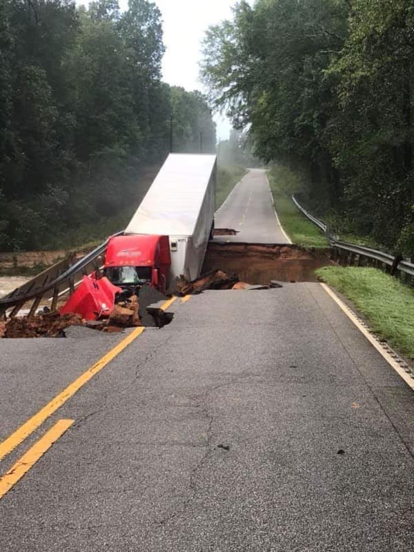 A catastrophic infrastructure breakdown and car fail where a massive red semi-truck with a white trailer has plunged nose-first into a giant sinkhole that completely split a rural asphalt highway.