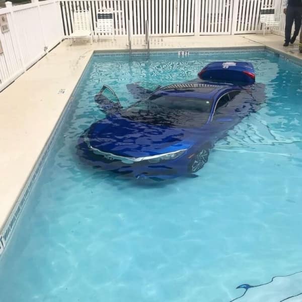 A bizarre residential car fail showing a modern bright blue sedan completely submerged at the bottom of a clear outdoor swimming pool with its trunk popped open.
