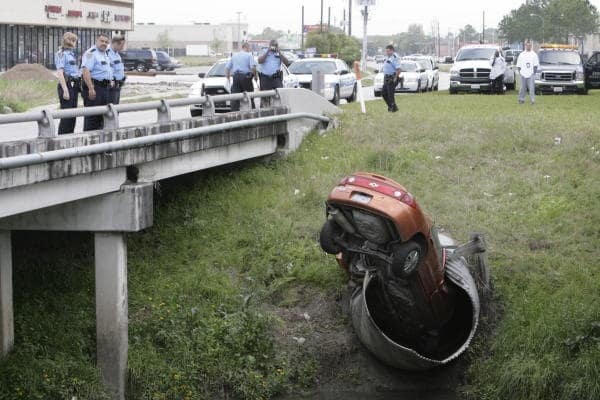 A spectacular drainage ditch car fail where an orange sedan is wedged perfectly upside down and nose-first inside a circular metal culvert pipe while police look on from a bridge above.