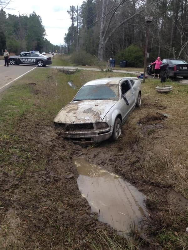 A muddy roadside ditch disaster highlighting a car fail where a silver Ford Mustang is wedged deep in a pool of brown sludge with a local sheriff's cruiser parked on the highway behind it.