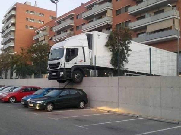 A high-altitude parking lot car fail captures a massive white semi-truck cab hanging precariously over a high concrete retaining wall, directly dangling above a line of parked compact vehicles below.