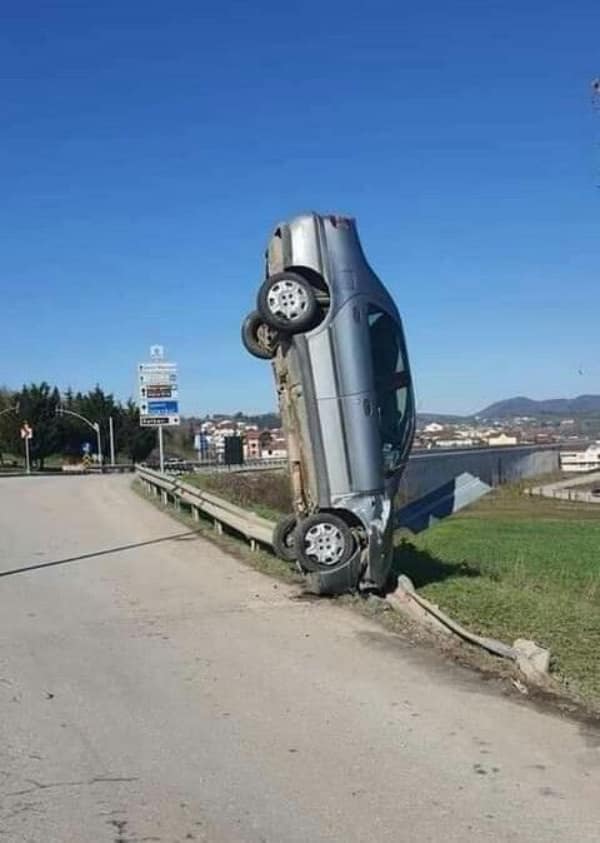 A physics-defying roadside car fail where a silver sedan is balanced completely vertically on its crushed front nose against a metal highway guardrail under a clear blue sky.