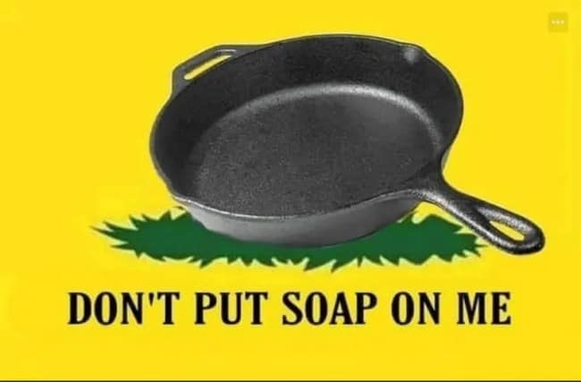 A political parody flag acting as a custom cast iron skillet meme, subverting the historic Gadsden "Don't Tread on Me" banner with an illustration of a black skillet over green grass alongside the text, "DON'T PUT SOAP ON ME."