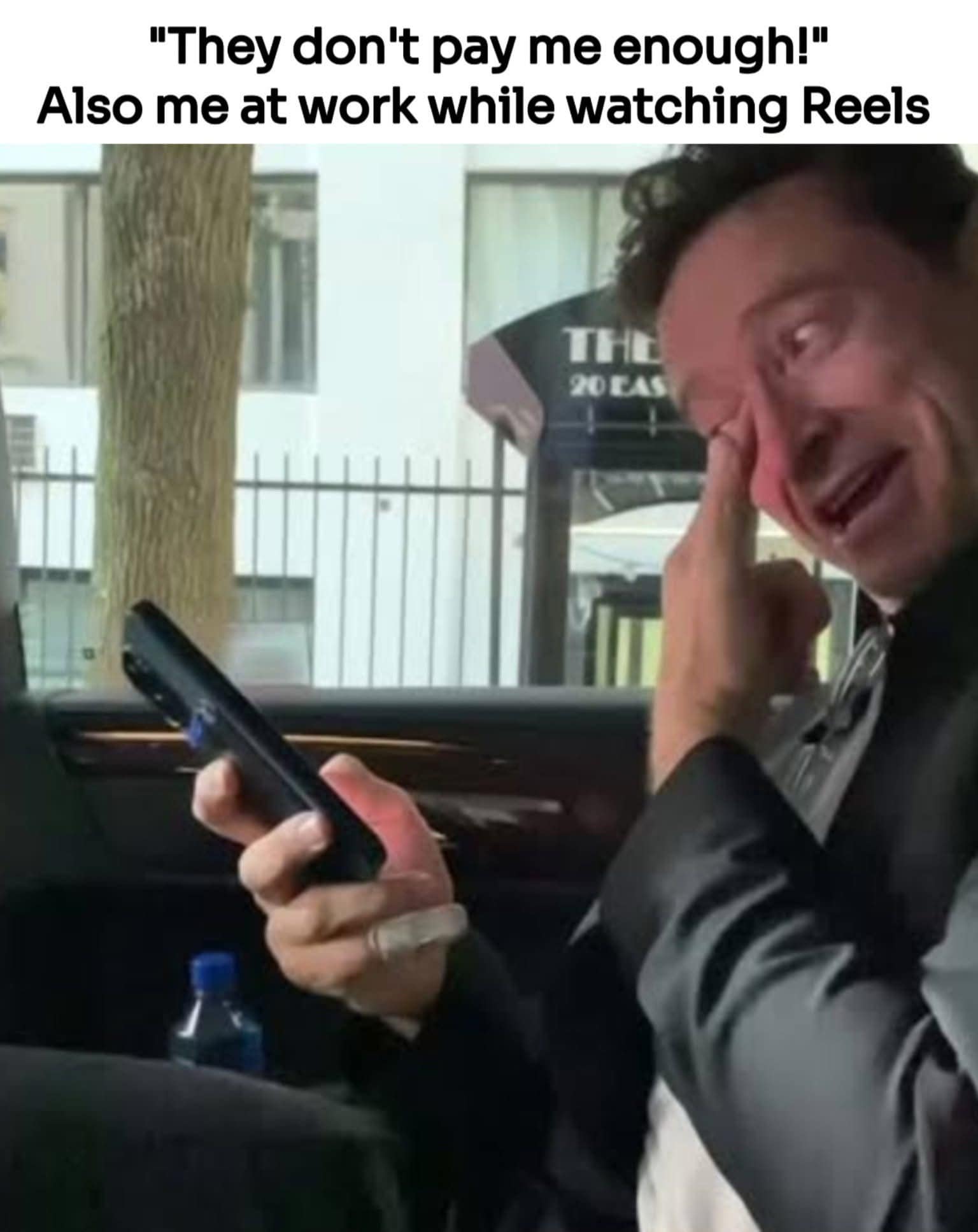 A universally relatable classic meme tackles workplace productivity with the top text, '"They don't pay me enough!" Also me at work while watching Reels.' The image features a candid close-up of actor Hugh Jackman sitting in the back of a vehicle, wiping a tear of laughter from his eye while completely absorbed by the screen of his smartphone.
