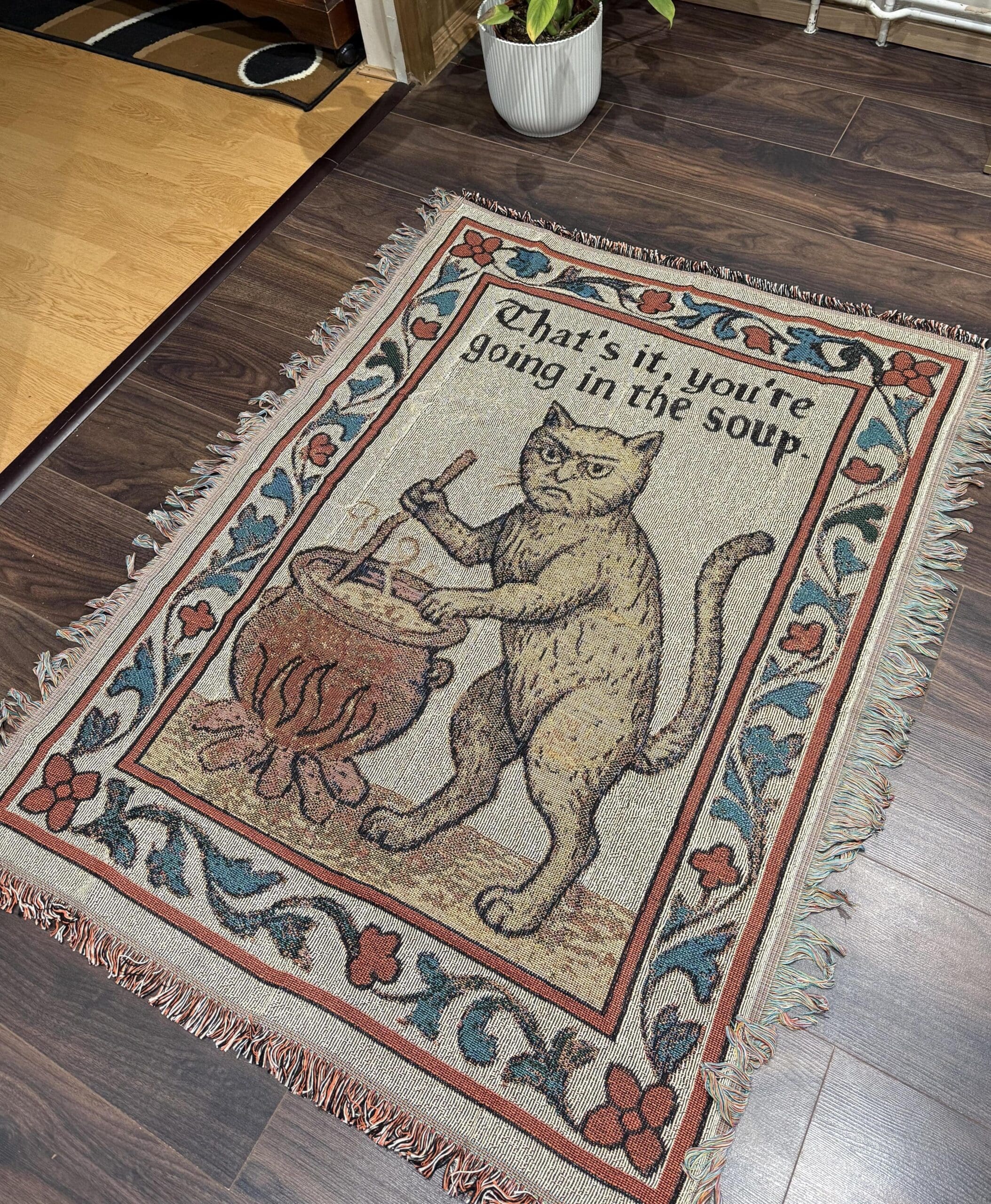 Woven tapestry rug on a wood floor depicting a medieval cat stirring a cauldron under the stitched threat text "That's it, you're going in the soup."