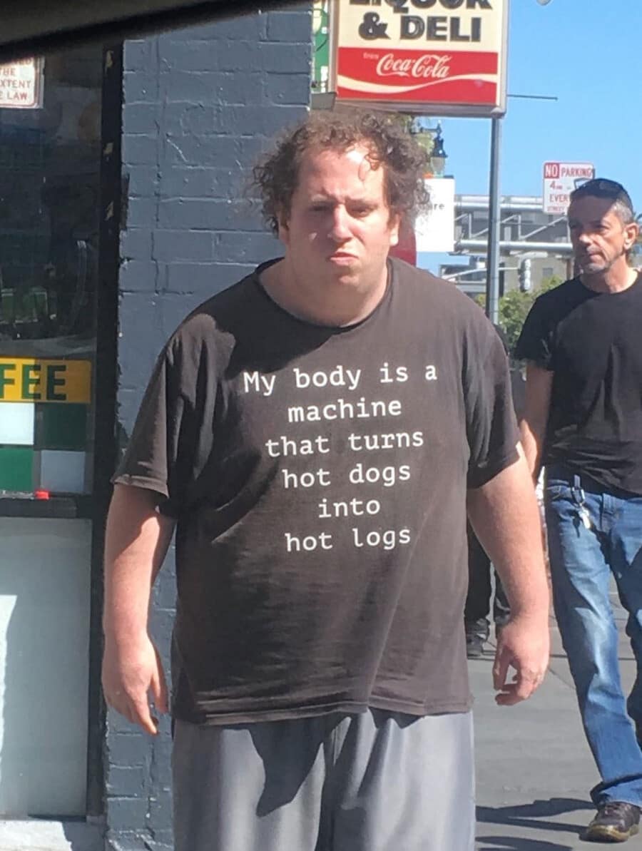 Man wearing a graphic t-shirt reading "My body is a machine that turns hot dogs into hot logs" on a city sidewalk in a classic meme.