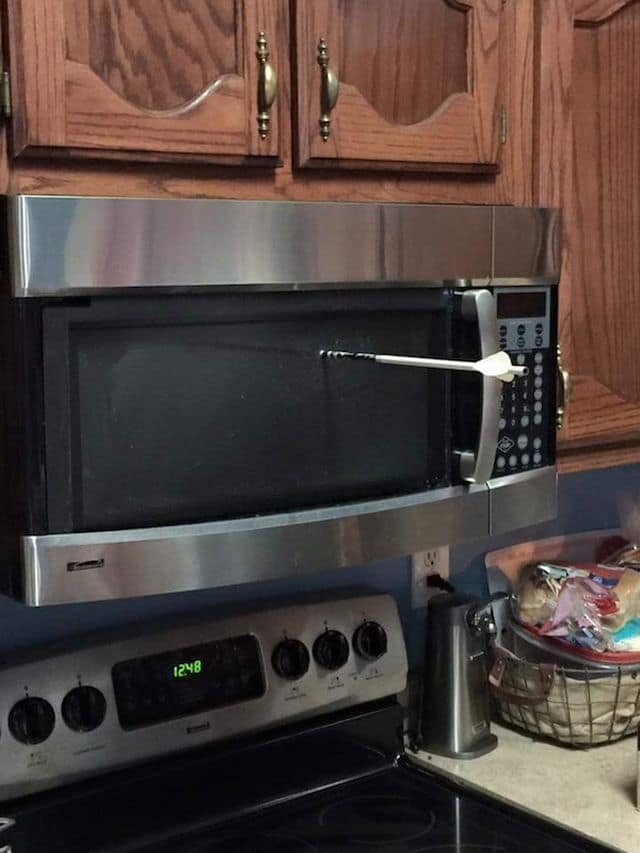 A classic meme visual displaying an unexplained household catastrophe where a target sport archery arrow has been shot cleanly through the dark glass door of a stainless steel microwave mounted over a stove.