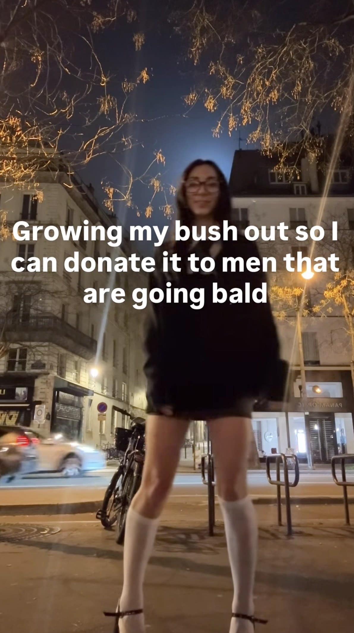 A viral street-vlog screenshot formatted as a classic meme, showing a girl standing under urban city trees at night with a wildly chaotic, literal hair-donation text overlay: "Growing my bush out so I can donate it to men that are going bald."