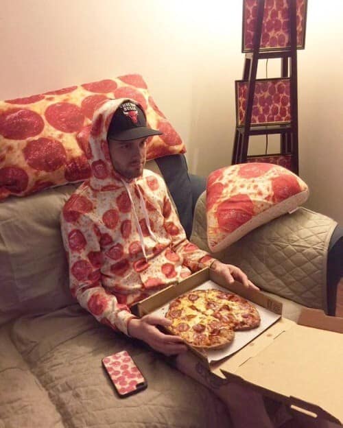 A man sits on a bed surrounded by pepperoni pizza-themed décor while wearing a pizza hoodie and holding an actual pizza box, creating a classic meme tableau of total commitment to the bit, the lifestyle, and possibly a marinara-based personality disorder.