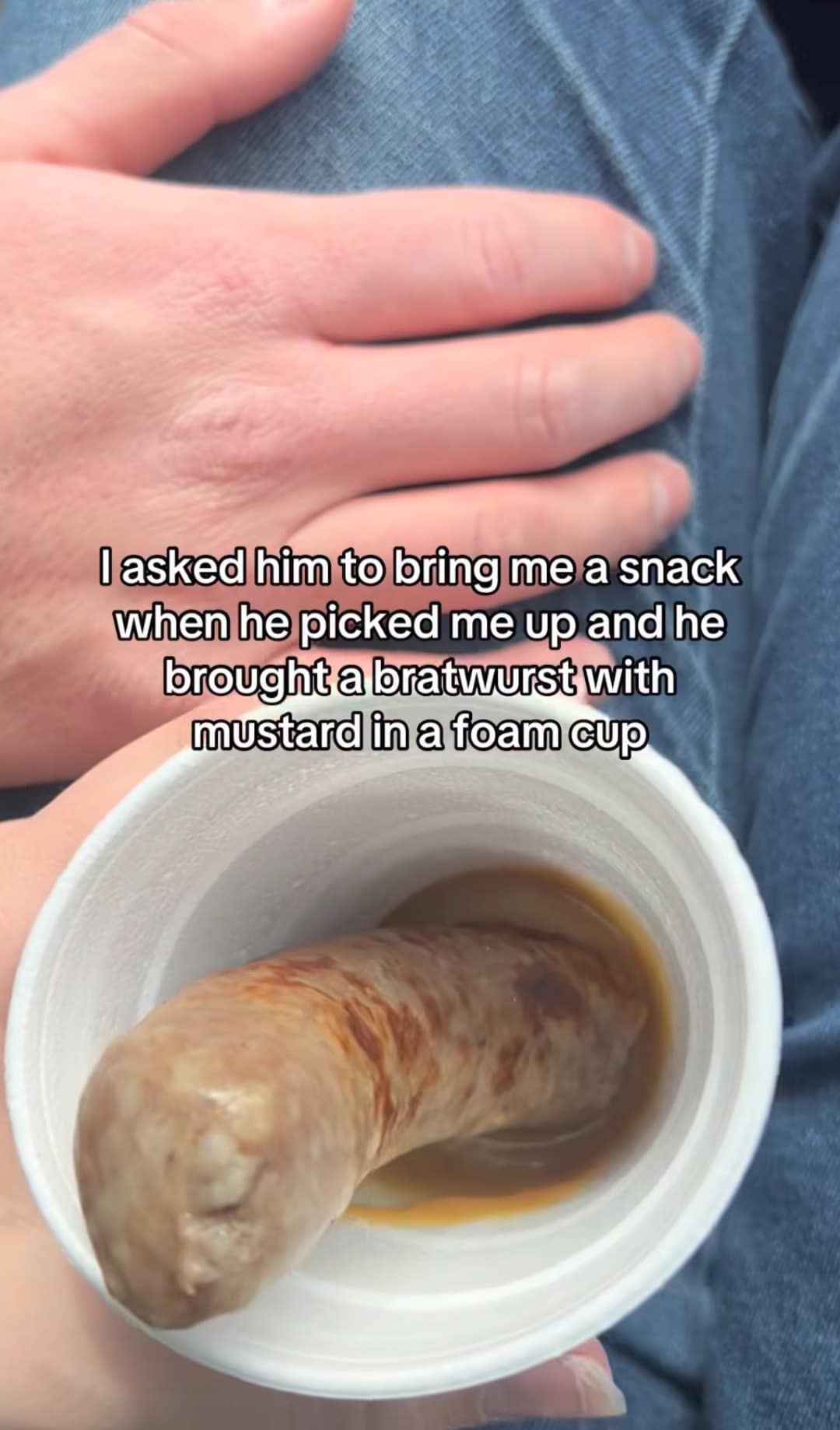 This chaotic relationship-themed classic meme captures a first-person perspective photo inside a moving car showcasing a truly unhinged drive-thru snack presentation. A passenger holds a white styrofoam coffee cup containing a single, large grilled bratwurst sausage drowned in a shallow puddle of brown mustard, highlighting a partner's hilariously literal interpretation of bringing back a snack.