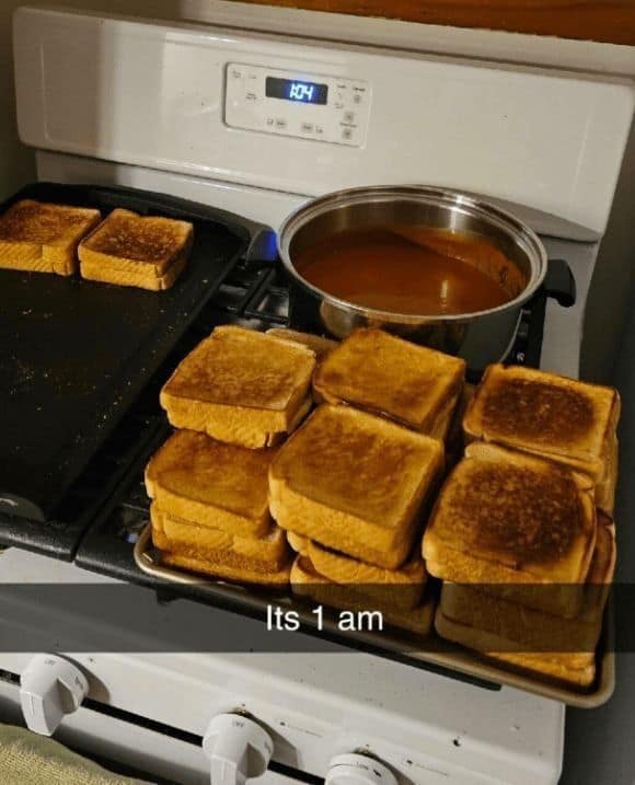A kitchen stovetop at 1:00 AM crowded with a massive, multi-tiered stack of golden grilled cheese sandwiches on a baking tray next to a bubbling pot of tomato soup in a late-night classic meme.