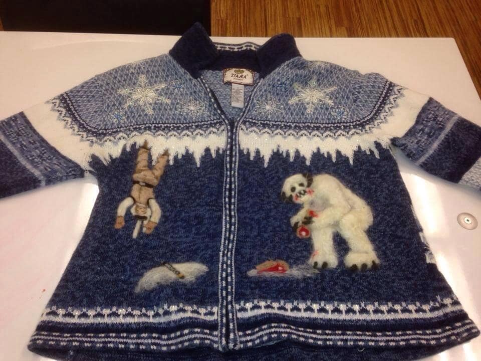 A fan-centric winter apparel photo circulating as a classic meme, showcasing a blue knit cardigan sweater designed with an intricate, custom scene from The Empire Strikes Back. The left side depicts Luke Skywalker hanging upside down in the frozen ceiling of the Wampa cave, while the right side features a plush, textured Wampa monster eating next to a bloody bone.