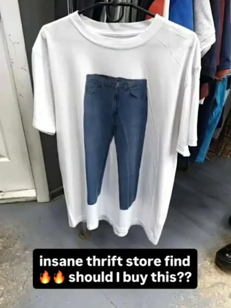 A clothing rack photo inside a secondhand thrift shop showing a plain white t-shirt with a highly realistic, full-sized graphic of straight-leg blue jeans printed directly onto the center torso, styled as a funny classic meme.