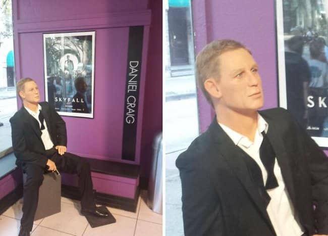 Awkward wax figure of Daniel Craig as James Bond sitting in a purple display.