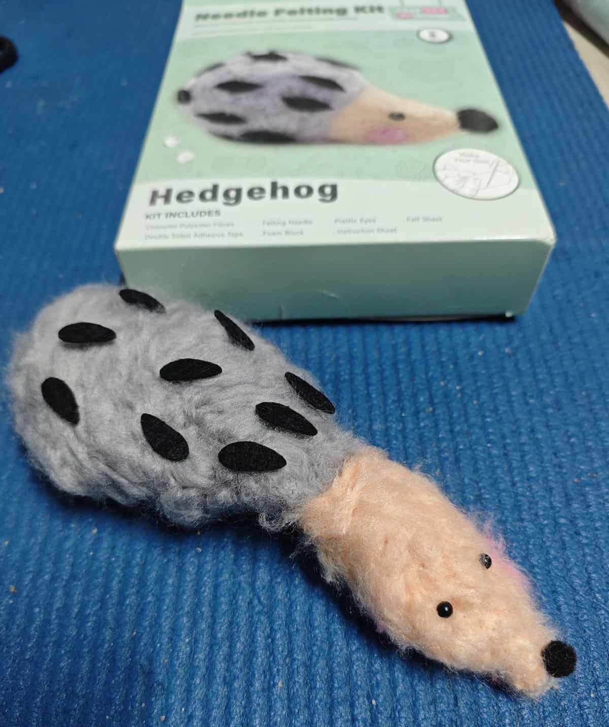 Elongated, misshapen gray and pink felt rodent creature lying in front of its retail hedgehog packaging, demonstrating a hilarious example of cursed crafts.