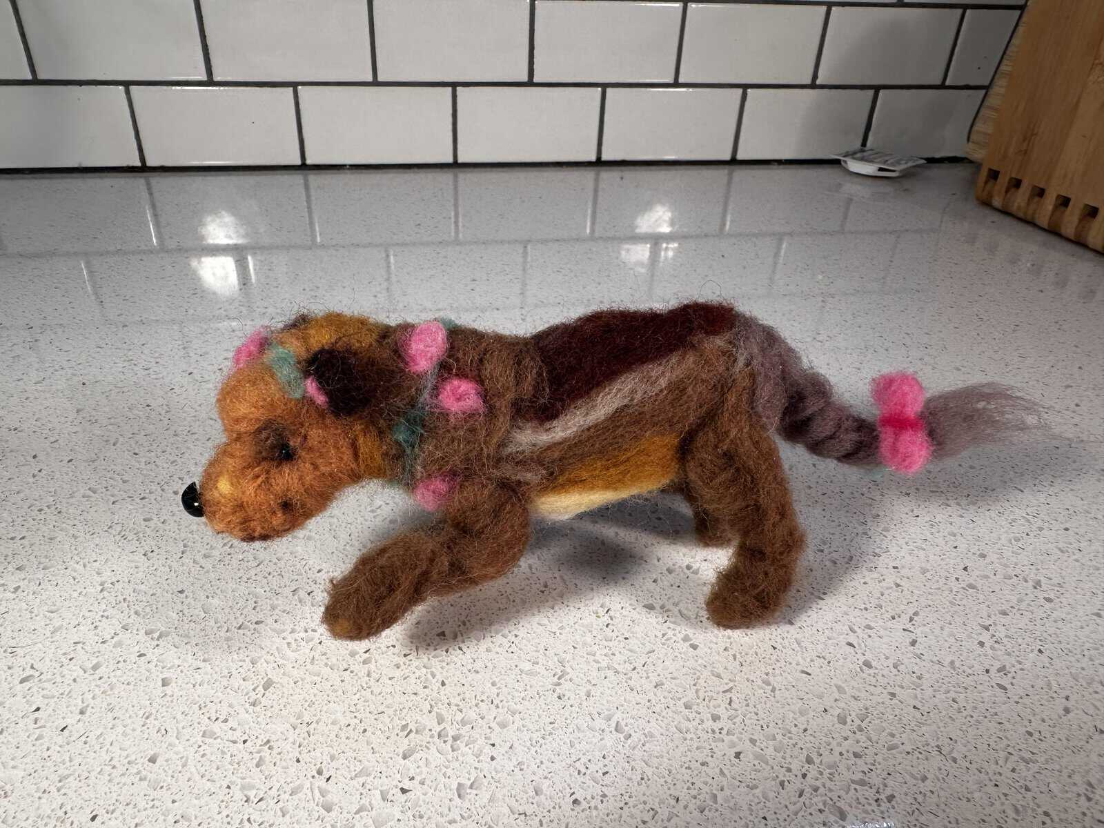A loosely sculpted, oddly proportioned brown and dark-brown felted quadruped creature with hot-pink wool bows tied on its ears, neck, and braided tail, standing on a kitchen counter as an example of cursed crafts.