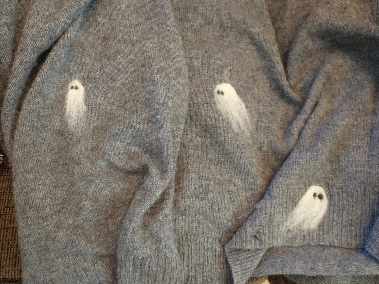 Close-up of three small, ghostly white spirits meticulously needle-felted directly onto the wool fabric of a grey knit cardigan sweater, adding an element of cozy, spooky style to funny felt art.
