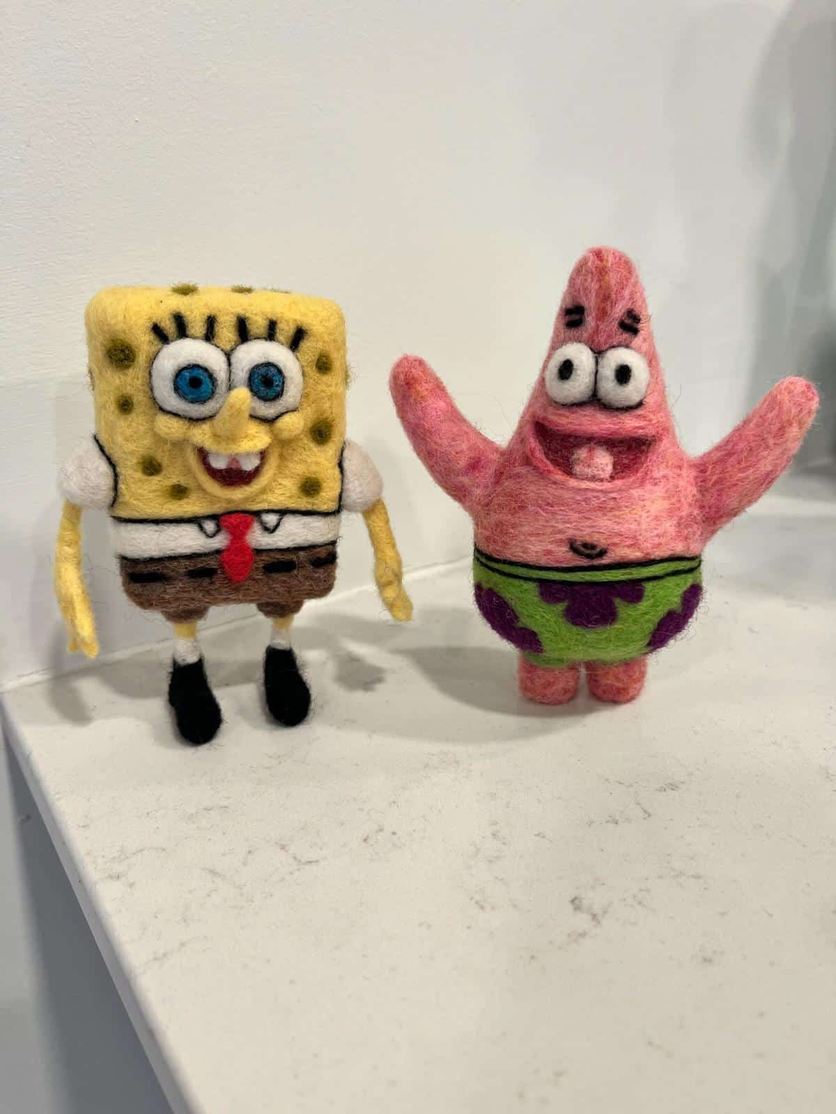 Clean, three-dimensional wool miniature figurines of SpongeBob SquarePants and Patrick Star standing side-by-side on a white kitchen counter surface, sculpted using precise funny felt art techniques.