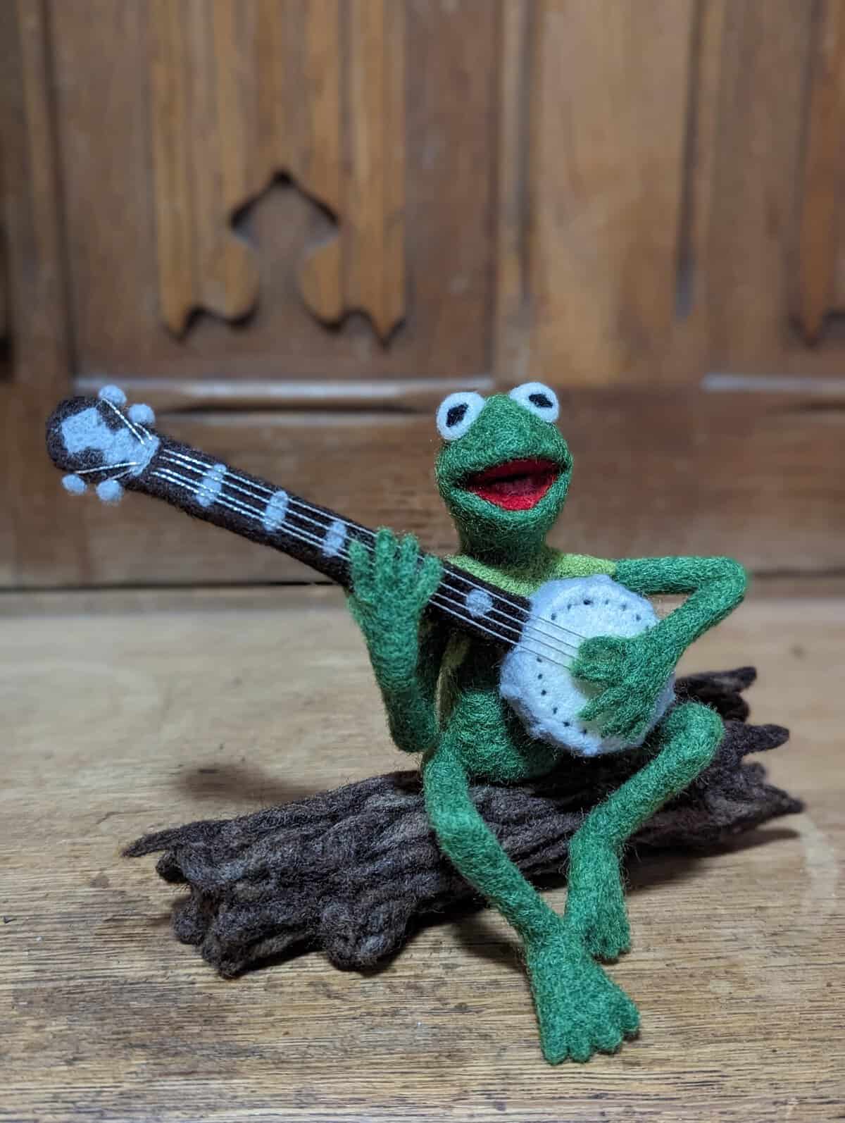 A wonderfully accurate needle-felted sculpture of Kermit the Frog sitting cross-legged on a dark brown textured wool log while happily playing a tiny, detailed white and black five-string banjo, captured as a musical tribute in funny felt art.