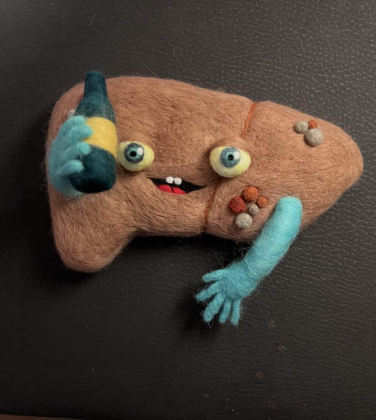 Goofy, buck-toothed human liver sculpture holding a beer bottle with cyan blue arms, showcasing a piece of darkly comedic and funny felt art.