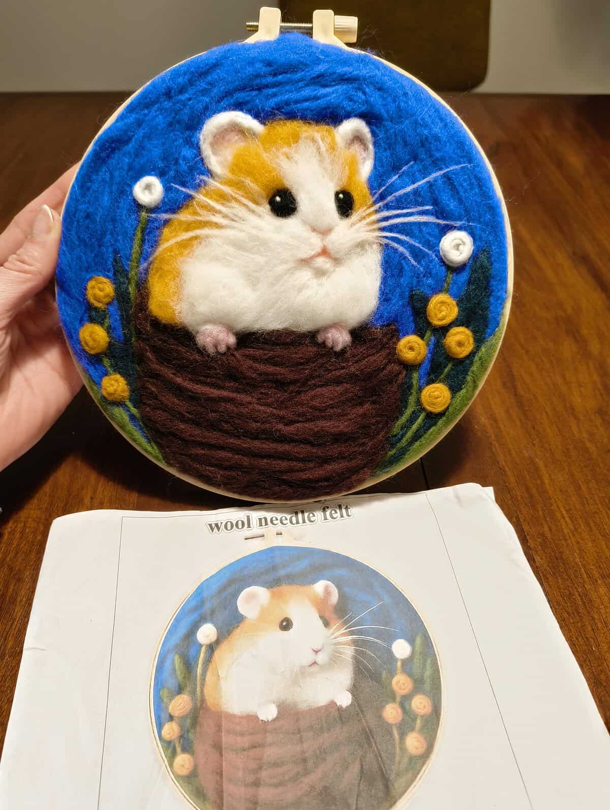 Circular embroidery hoop containing a three-dimensional wool needle felted hamster popping out of a brown basket, comparing the final product to a funny felt art template.