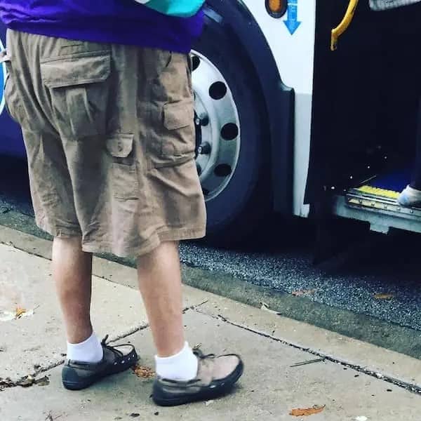 Close-up of a person standing on a sidewalk wearing baggy brown cargo shorts and grey slip-on shoes with white socks.