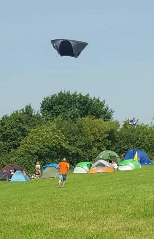 A black camping tent floating high in the sky above a grassy campsite filled with other tents.