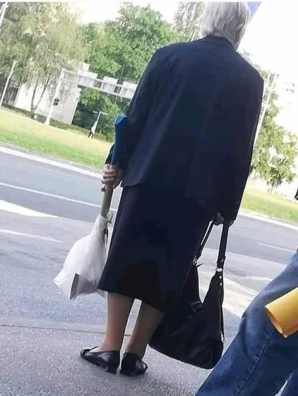 Back view of an older woman holding a crutch with a large ax hidden inside a plastic bag.