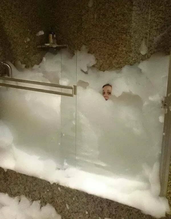 A person's face barely visible peeking out from a glass shower stall completely filled with soap suds.