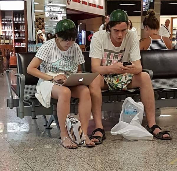 Two passengers sitting in an airport terminal waiting area wearing hollowed-out watermelon halves as helmets.