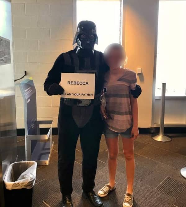 A dad dressed in a full Darth Vader costume holding a sign that reads Rebecca I am your father.