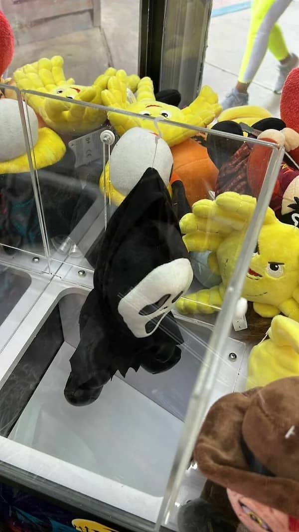 Ghostface plush toy hanging sadly by its hood inside an amusement arcade claw machine.