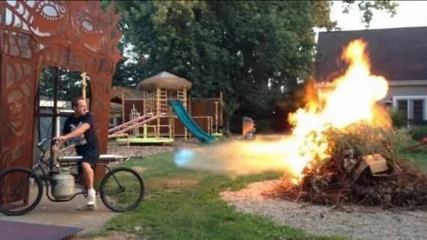 Backyard photo of a man riding a custom propane tank bicycle that acts as a powerful flamethrower.