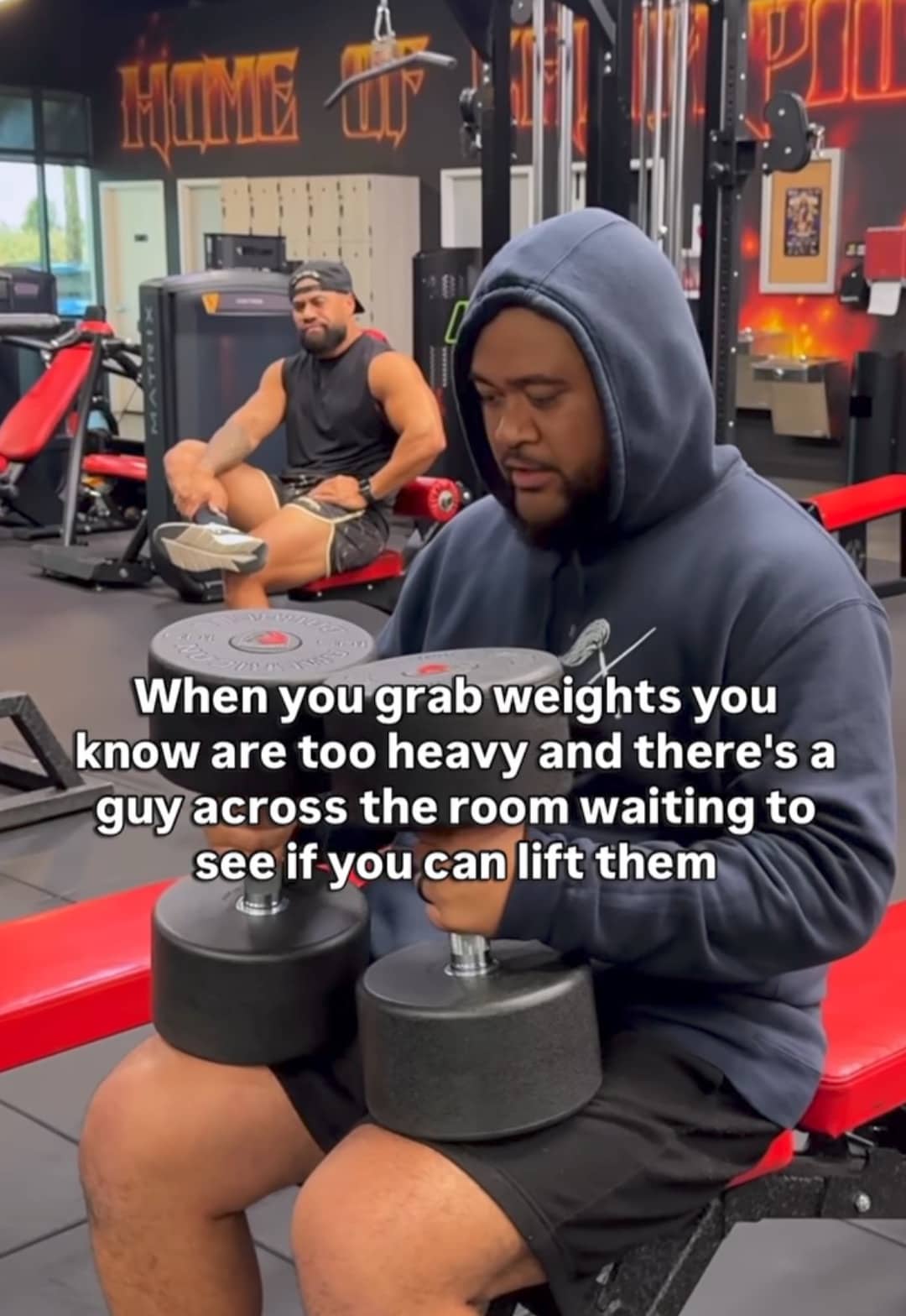 Man in hoodie holding heavy dumbbells in gym with someone watching him lift meme.