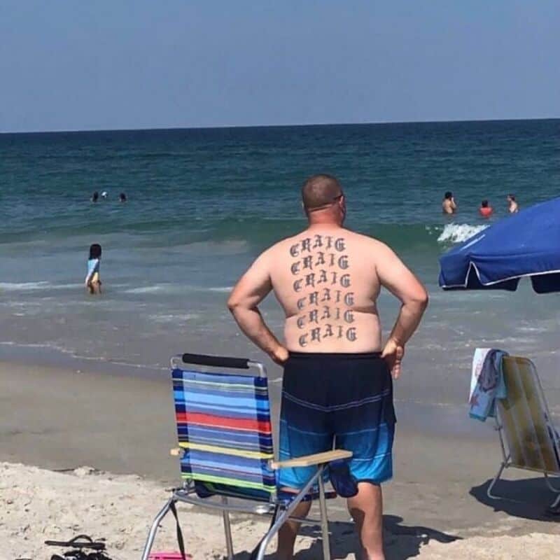 Shirtless man standing on a beach with the name Craig tattooed repeatedly down his back.